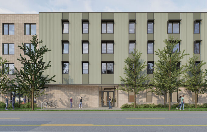 Modern multi-story building with a mix of light-colored brick and green paneling. Large windows are evenly spaced across the facade. The entrance is centrally located and surrounded by trees and shrubs. Pedestrians and cyclists can be seen in front of the building, indicating an active environment.