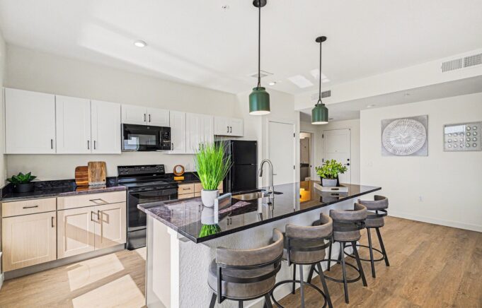 Modern kitchen with a black granite island, bar stools, and hanging lights. Light wood cabinets, black appliances, and green plants add a fresh touch.