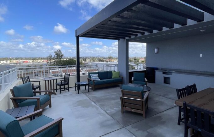 A rooftop terrace with comfortable seating including chairs and couches, a dining table, and an outdoor kitchen area. The sky is partly cloudy, and the view overlooks a cityscape with buildings in the distance. The space is designed for relaxation and socializing.