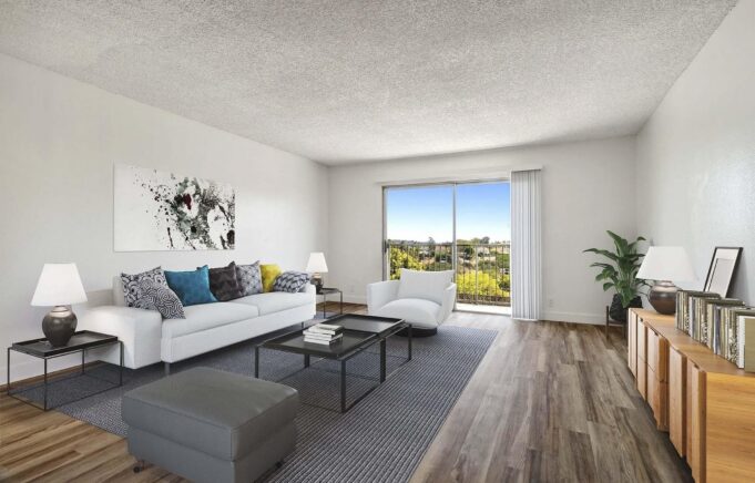 A modern living room featuring a white sofa with colorful throw pillows, a glass coffee table, and a light gray area rug. Two lamps are on either side of the sofa. A stylish cabinet holds books, and a large window provides a view of greenery outside. The walls are painted white and the floor has wooden planks
