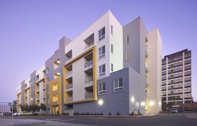 Modern apartment building at dusk, showcasing a contemporary design with multiple balconies and large windows. The building features a mix of gray and yellow accents, along with surrounding parking and green landscaping. In the background, another high-rise building is visible.